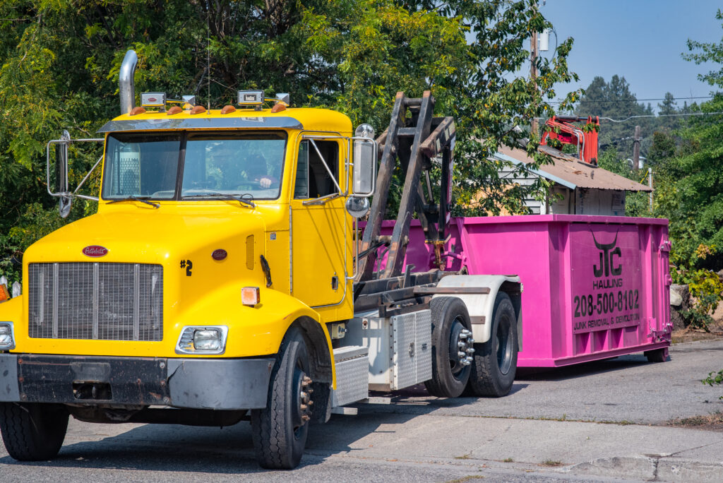 Yellow truck from JTC Demolition delivering a pink dumpster for waste disposal in Spokane, surrounded by greenery and residential buildings.