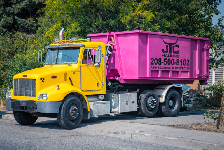 Yellow dumpster truck with pink container labeled "JTC Hauling 208-500-8102 Junk Removal & Demolition" parked on a residential street in Spokane Valley, surrounded by greenery.
